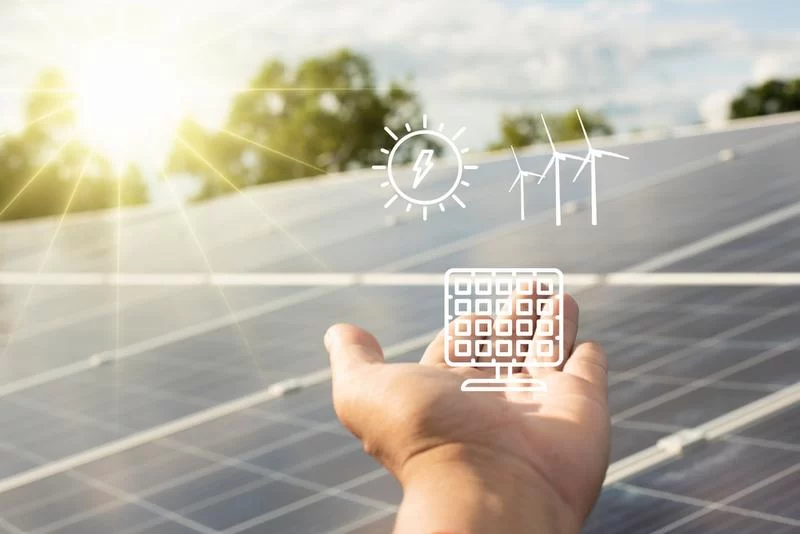 A hand holds a solar panel with a wind turbine in the background, representing innovative solar energy concept